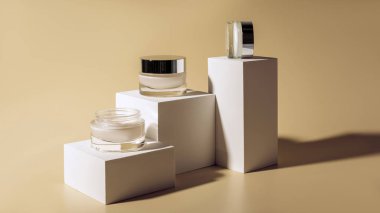 close up view of facial and body creams in glass jars on white cubes on beige background clipart