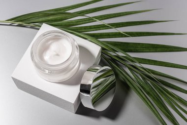 top view of moisturizing cream in glass jar with palm leaf on grey background clipart