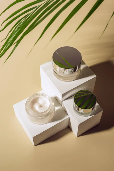 在米色背景上的白色立方体上的玻璃罐子上的绿色棕榈叶和体霜的特写视图 — 图库照片
