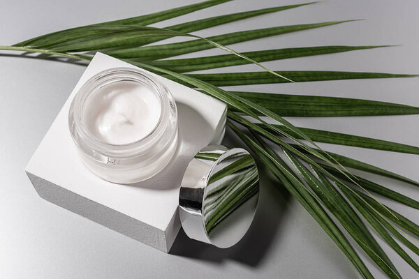 top view of moisturizing cream in glass jar with palm leaf on grey background