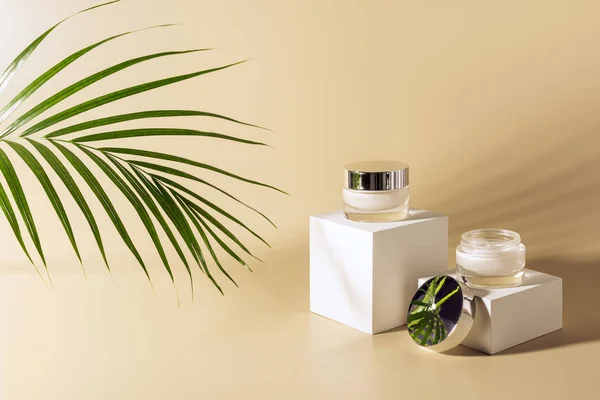 Close up view of green palm leaf, facial and body creams in glass jars on white cubes on beige background — Stock Image Close View Green Palm Leaf Facial Body Creams Glass Jars Stock Picture