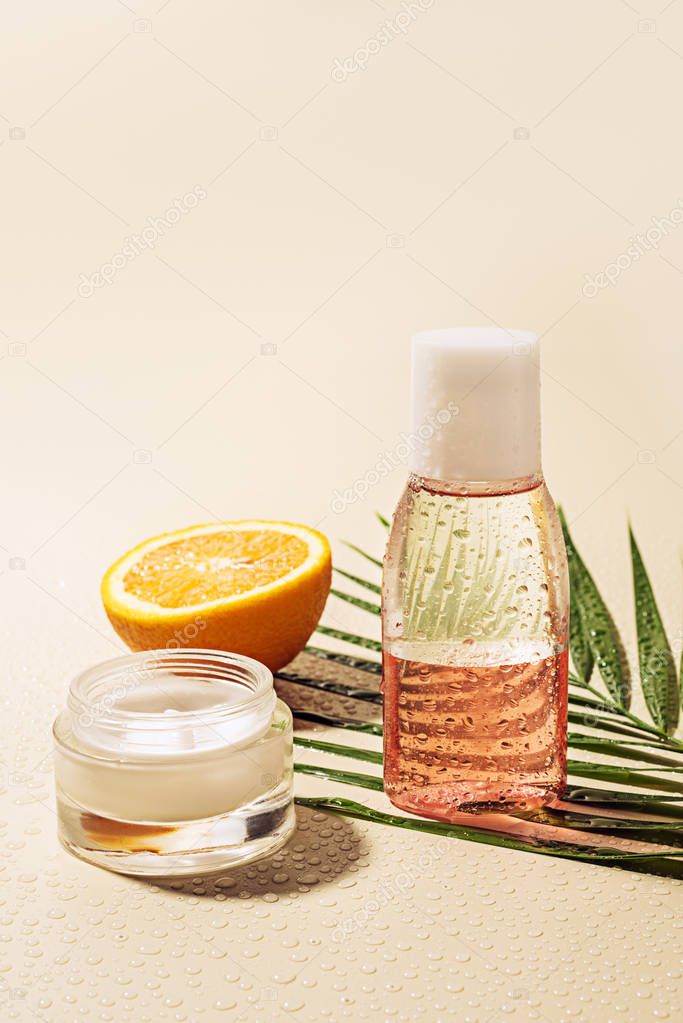 close up view of facial cream and lotion, palm leaf and orange half with water drops on beige background