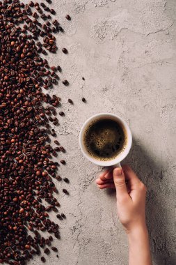 cropped shot of woman holding cup of coffee on concrete surface with spilled coffee beans clipart