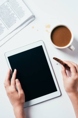 cropped shot of woman using tablet while eating chocolate chip cookie with coffee on white surface clipart