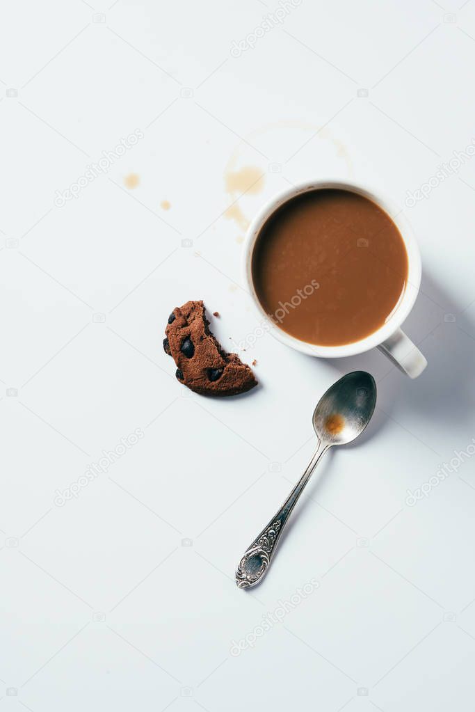top view of cup of coffee with bitten chocolate chip cookie and spoon on white surface