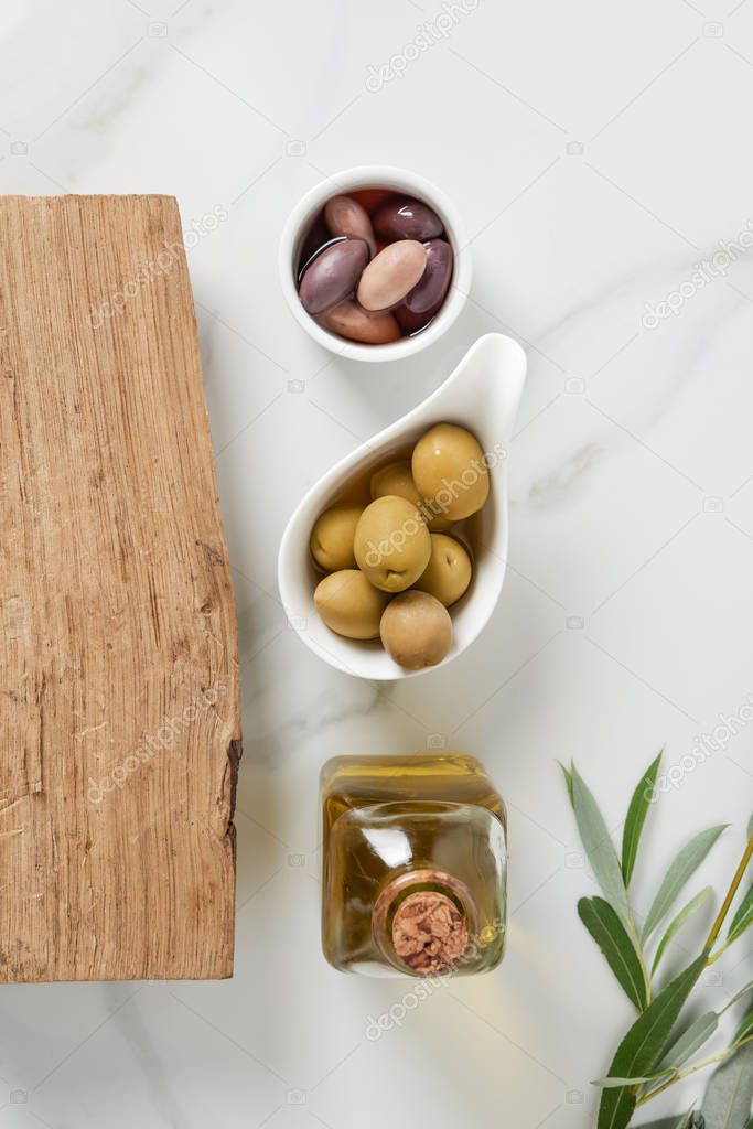 top view of bottle of olive oil, log and olives in bowls on marble table 