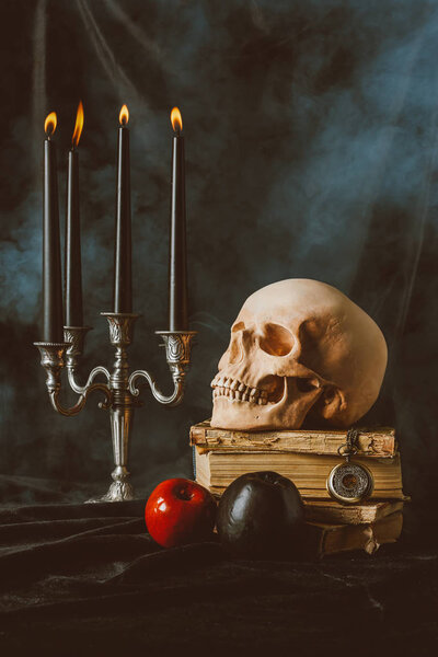 канделябр, череп на Хэллоуин, древние книги с черными и красными яблоками на черной ткани с дымом
