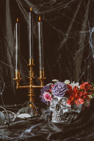 hourglass, silver skull with flowers and candelabrum with candles on black cloth with spider web, decorations for halloween