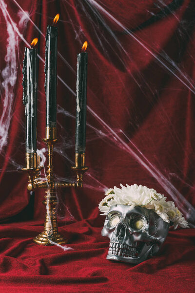 серебряный череп с цветами и канделябр со свечами на красной ткани с паутиной
 