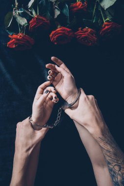 cropped shot of man holding female hands in handcuffs above black fabric with red roses clipart
