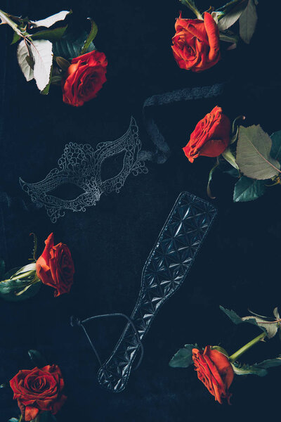 top view of lace mask, leather spanking paddle and beautiful red roses on black