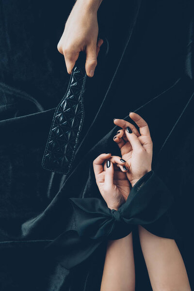 cropped shot of woman with tied hands and man holding black leather spanking paddle