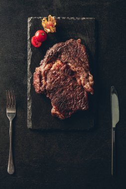 top view of cooked steak and cherry tomato on black wooden board, knife and fork on table in kitchen