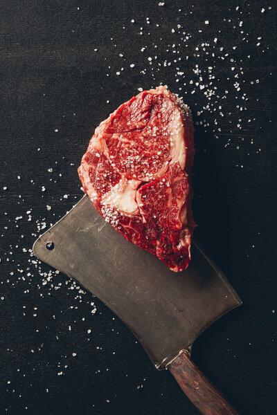 top view of raw meat steak, salt and cleaver on tabletop in kitchen