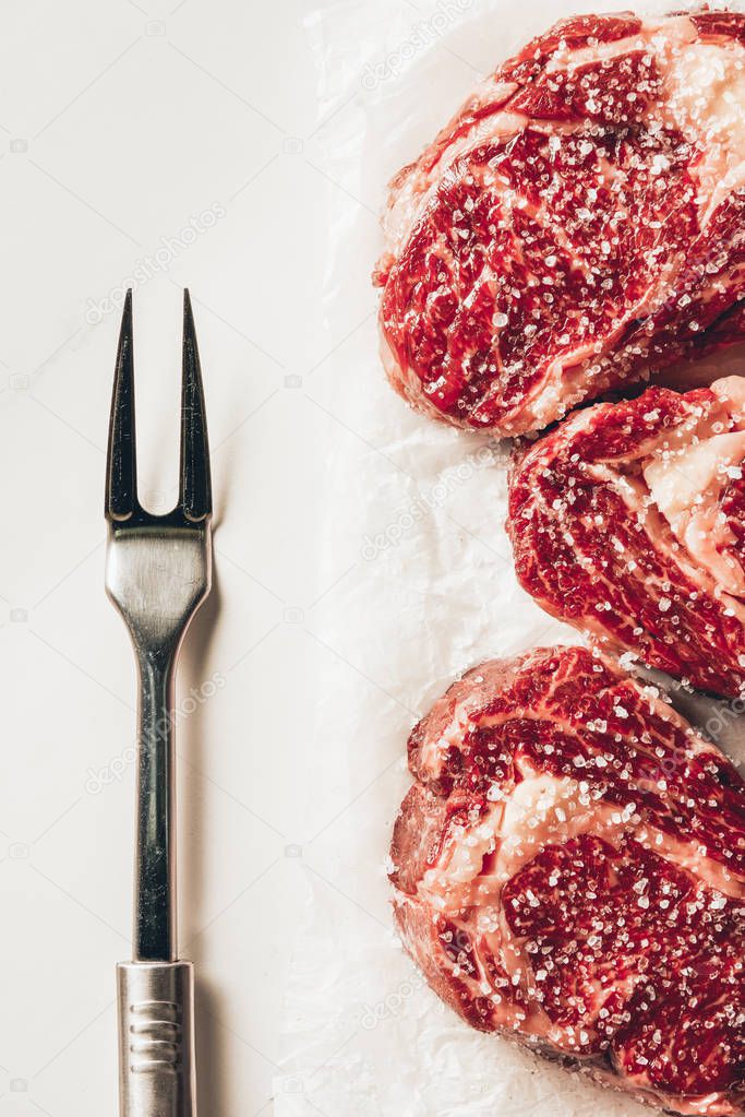elevated view of three pieces of raw meat steaks and meat fork on table in kitchen