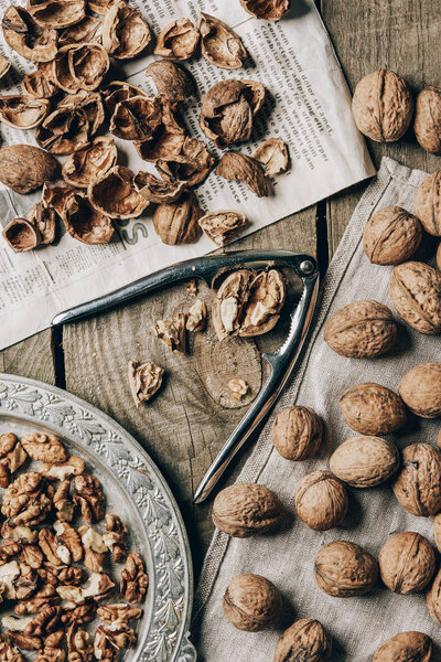 top view of whole and cracked walnuts, nutcracker, vintage plate, cloth and newspaper on wooden table