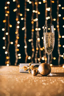 glass of champagne, golden baubles and present on garland light background, christmas concept clipart