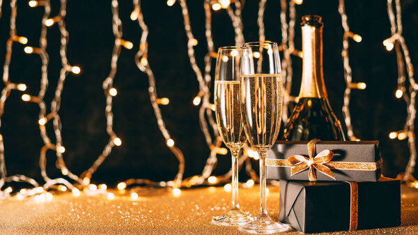 two glasses of champagne, presents and bottle on garland light background, christmas concept