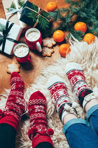 cropped shot of women in winter socks at background with wrapped christmas presents and cups of hot chocolate with marshmallows