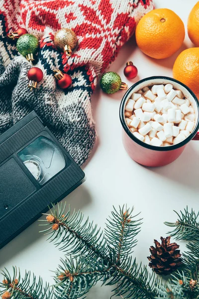 top view of video cassette, sweater, christmas balls and cup of hot drink with marshmallows on white surface