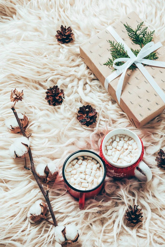 flat lay with cup of cocoa with marshmallows, wrapped christmas present and cotton twig on woolen background