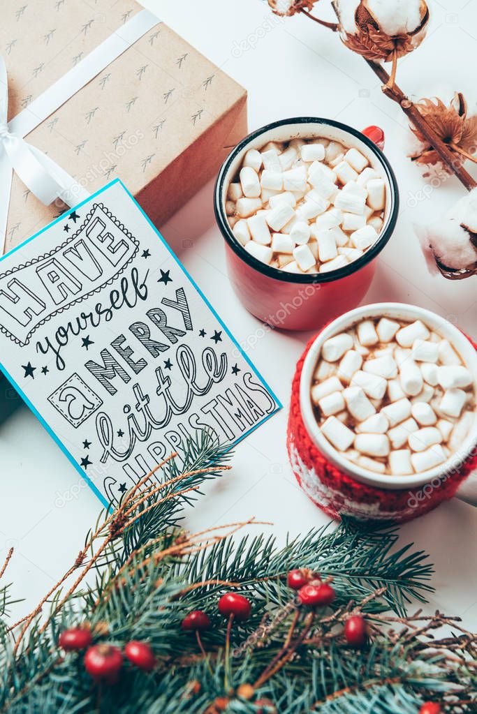 top view of postcard with have yourself marry little christmas lettering, present and cups of cocoa drinks with marshmallows on white tabletop