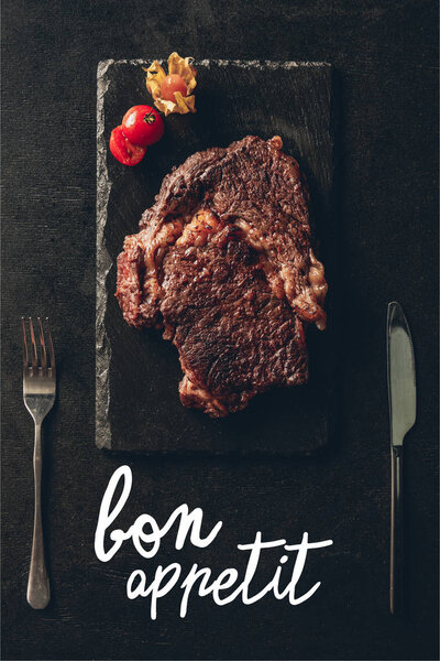 top view of cooked steak and cherry tomato on black wooden board, knife and fork on table in kitchen, bon appetit lettering