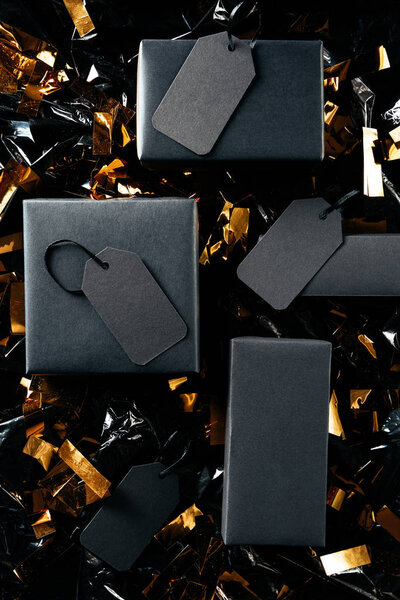 flat lay with black boxes with blank price tags and golden confetti on black wrapping paper backdrop, black friday concept