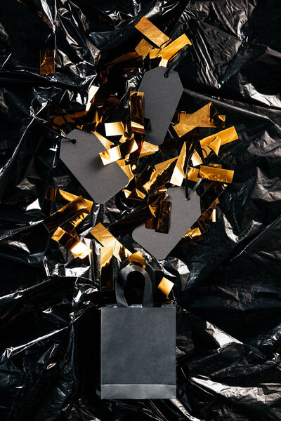 top view of paper shopping bag with blank price tags and golden confetti on black wrapping paper backdrop, black friday concept