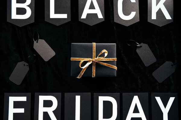 top view of black friday lettering, wrapped gift and blank price tags on black backdrop