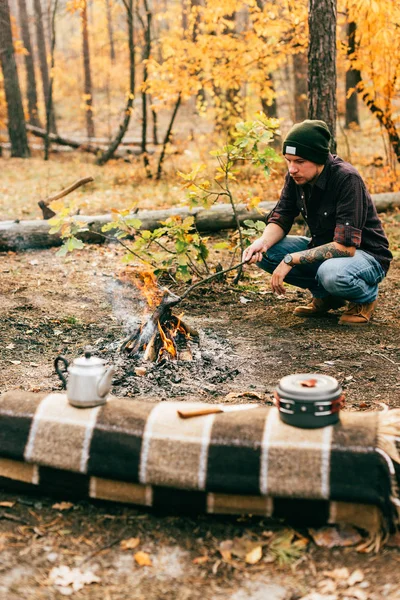 adult man cooking food by fire outdoors in autumn forest