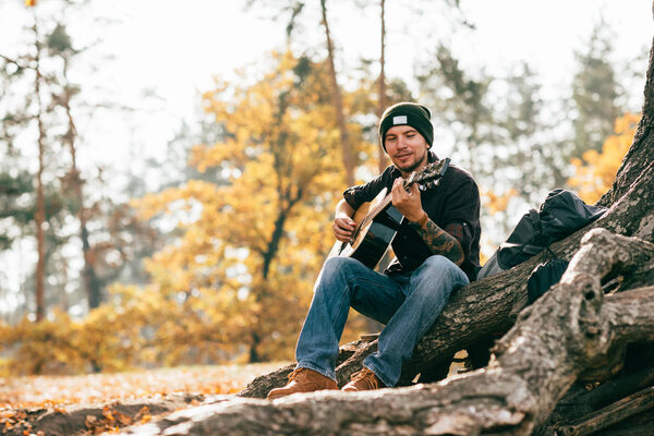 взрослый мужчина играет на акустической гитаре, сидя на дереве в парке
 