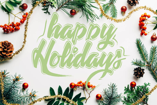 flat lay with festive arrangement of pine tree branches, common sea buckthorn and christmas decorations on white tabletop with "happy holidays" lettering