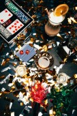 erhöhter Blick auf alkoholische Cocktails, Spielkarten, Pokerchips und Partyhörner auf einem mit goldenem Konfetti bedeckten Tisch 