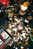 Draufsicht auf alkoholische Cocktails, Spielkarten, Pokerchips und Partyhörner auf einem mit goldenem Konfetti bedeckten Tisch 