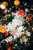 erhöhter Blick auf Spielkarten, alkoholische Cocktails mit Orangenscheiben, Whiskey und Partyhörner auf einem mit goldenem Konfetti bedeckten Tisch 