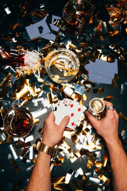partial view of man with playing cards and alcohol sitting at table covered by golden confetti clipart