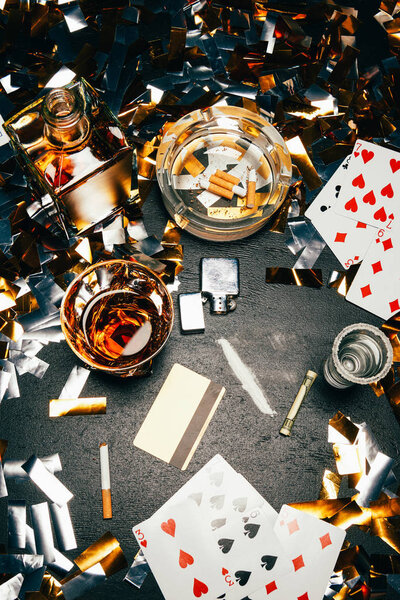 view from above of playing cards, cigarettes, whiskey, rolled banknote, credit card and cocaine on table covered by golden confetti 