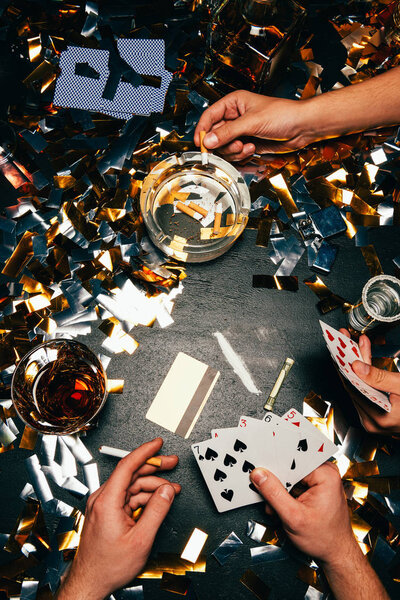 partial view of men playing poker with cocaine, alcohol and cigarettes at table covered by golden confetti