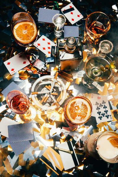 top view of cigarettes, alcoholic cocktails, playing cards, rolled banknote, credit cards and cocaine on table covered by golden confetti 