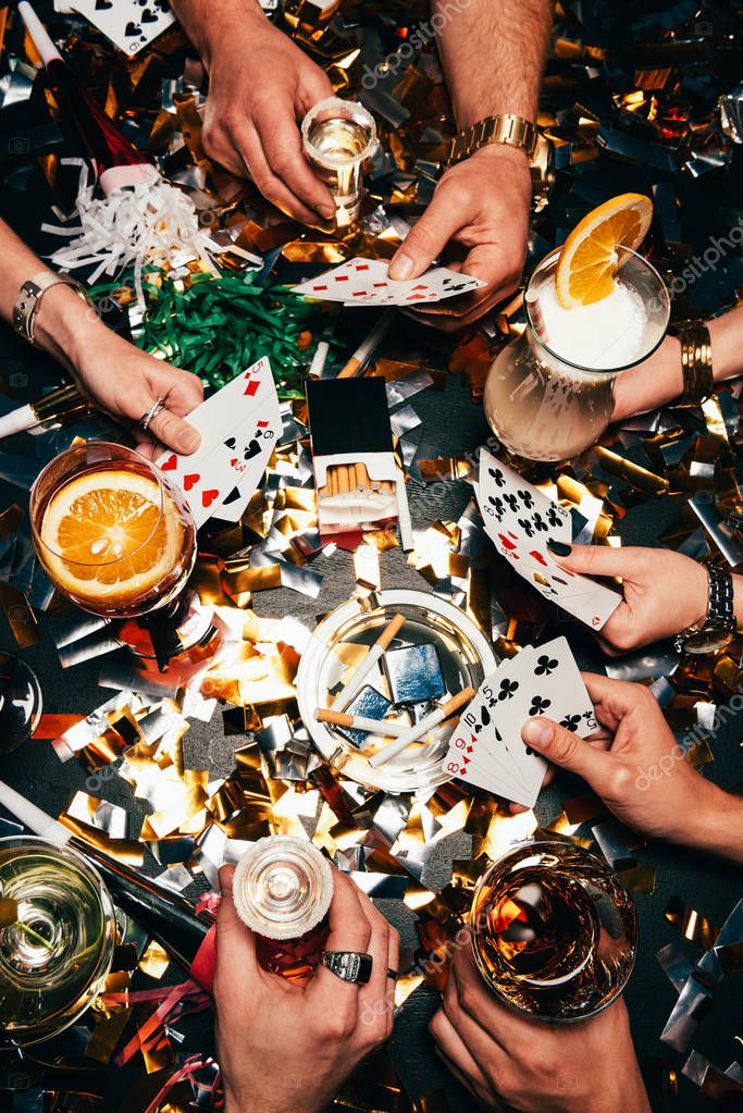 Cropped image of friends with alcoholic cocktails playing poker at table covered by golden confetti