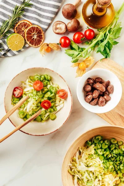 top view of salad on white bowl with bottle of oil, cherry tomatoes, lime and rosemary on surface 