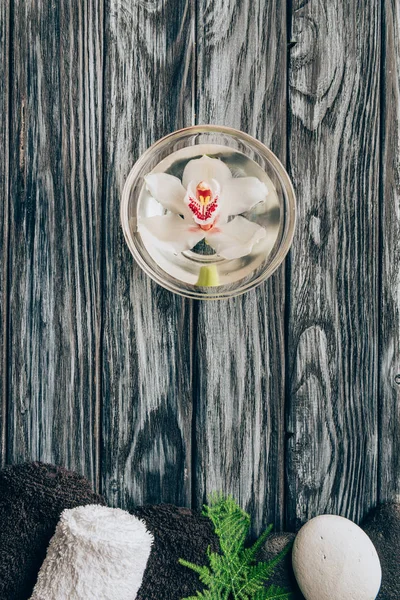 Vista superior de la flor de orquídea arreglada en un tazón, toallas y guijarros para spa y masaje en la mesa de madera - foto de stock