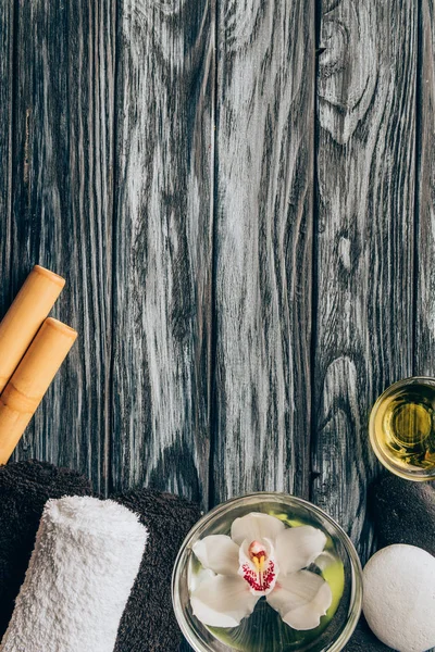 Vista dall'alto di asciugamani disposti, fiore di orchidea in ciotola, olio e bastoncini di bambù per spa e massaggio su sfondo di legno — Foto stock