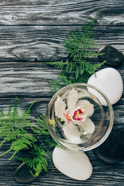 Disposición plana con spa y tratamiento de masaje con helecho, guijarros y flor de orquídea en cuenco sobre mesa de madera - foto de stock