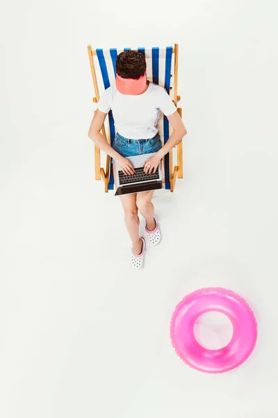 Vista aérea de la mujer sentada en una silla de playa a rayas y utilizando un ordenador portátil aislado en blanco - foto de stock