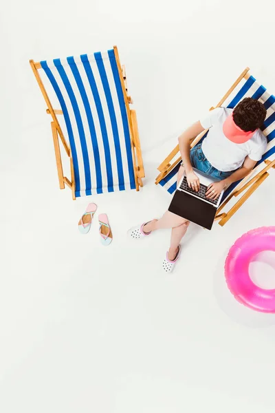 Vista aérea da mulher sentada na cadeira de praia e usando laptop isolado no branco — Fotografia de Stock