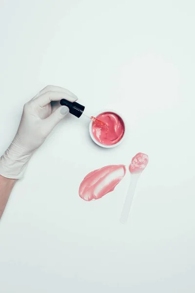 Uma mão em luva de látex segurando tubo perto do recipiente com máscara rosa, colher e mancha na superfície branca — Fotografia de Stock