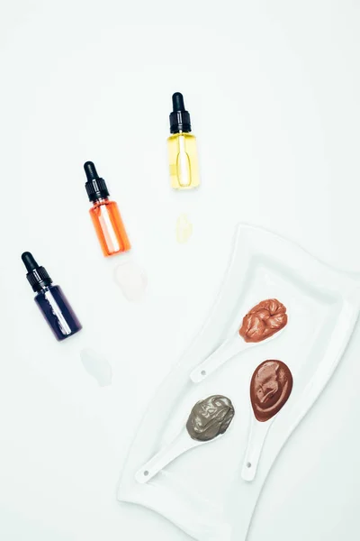 Vista superior de polvo de arcilla, botellas de cosméticos con tuberías, placa y cucharas con varias máscaras de arcilla de colores aislados en la superficie blanca - foto de stock