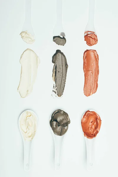 Leigos planos com colheres e várias manchas de máscara de argila coloridas colocadas em linha isoladas na superfície branca — Fotografia de Stock
